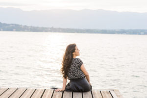 Woman sits on a dock looking away from camera water and mountains in the background